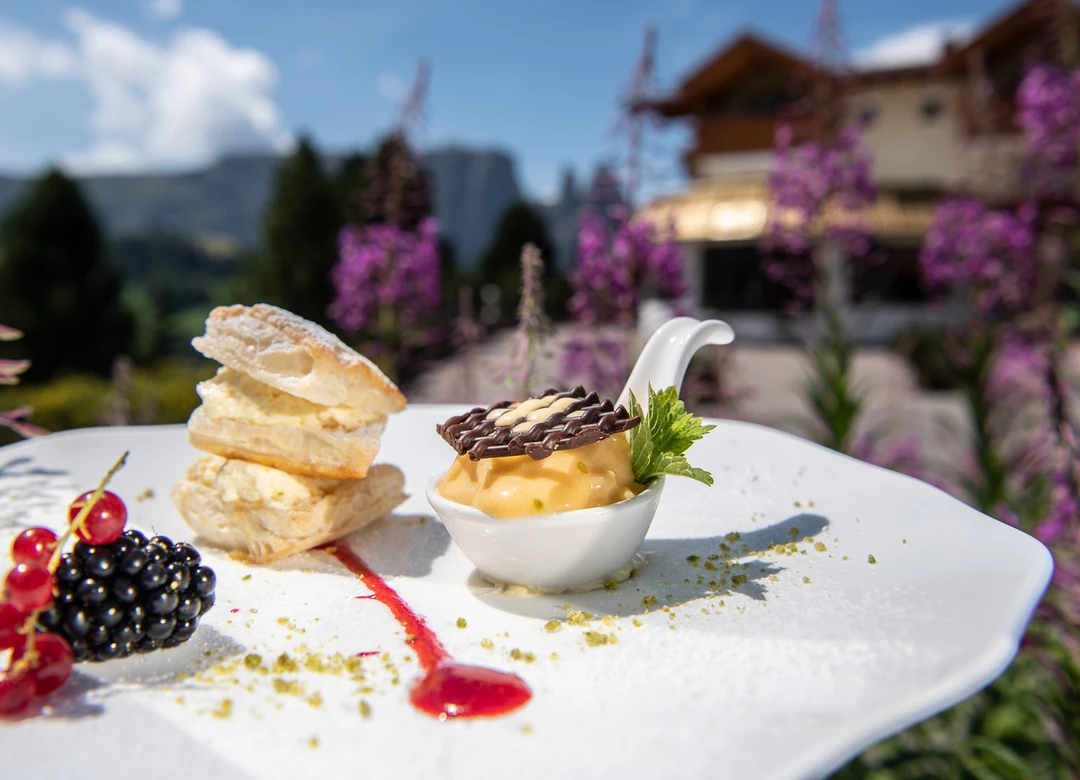 Un dessert delicato con pasta sfoglia, gelato e frutti di bosco su un piatto davanti a un paesaggio montano con fiori viola e un hotel sullo sfondo. - Bild größer darstellen