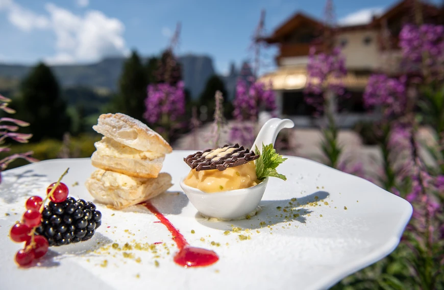 Un dessert delicato con pasta sfoglia, gelato e frutti di bosco su un piatto davanti a un paesaggio montano con fiori viola e un hotel sullo sfondo. - Bild größer darstellen