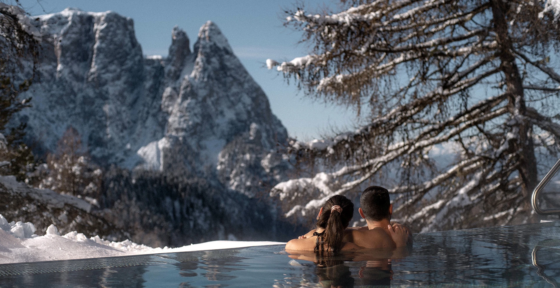 Una coppia si gode la piscina Infinity con vista sulle Dolomiti innevate e sul pittoresco paesaggio invernale dell'Alpe di Siusi sotto il sole splendente.