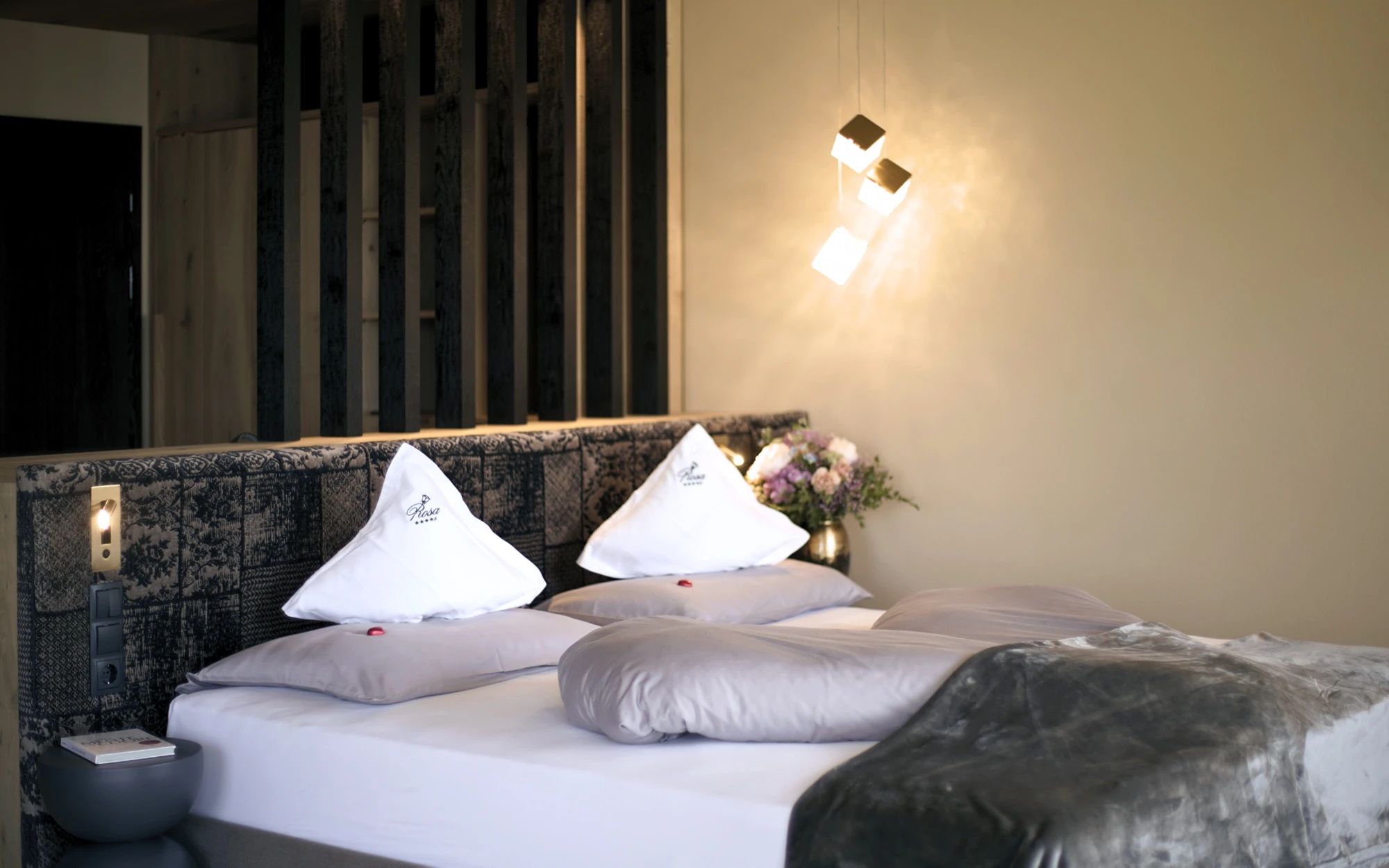 Un'accogliente camera d'hotel con letto matrimoniale, cuscini bianchi, coperte grigie e una moderna illuminazione calda creata da cubi luminosi sospesi.