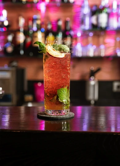 Un cocktail rinfrescante all'Hotel Rosa con vista sulle Dolomiti e sull'Alpe di Siusi, guarnito con menta e una fetta di mela davanti allo sfondo del bar. - Bild größer darstellen