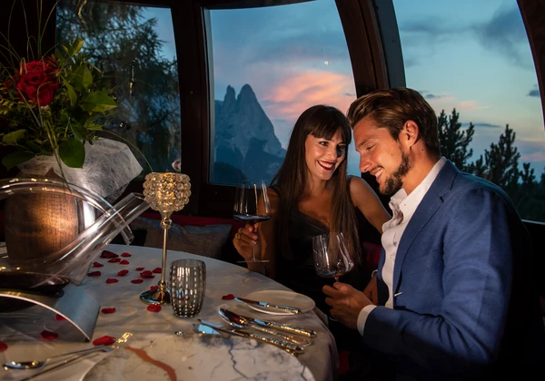 Una coppia si gode una cena romantica all'Hotel Rosa con vista sull'imponente paesaggio montano delle Dolomiti e sull'Alpe di Siusi al crepuscolo. - Bild größer darstellen