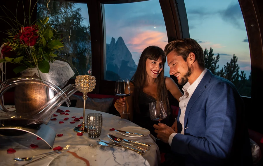 Una coppia si gode una cena romantica all'Hotel Rosa con vista sull'imponente paesaggio montano delle Dolomiti e sull'Alpe di Siusi al crepuscolo. - Bild größer darstellen