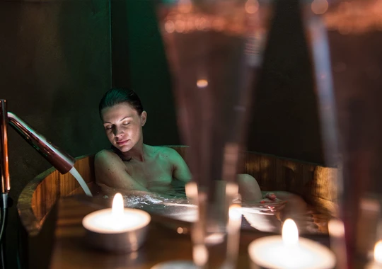Una donna si rilassa nella vasca idromassaggio in legno dell'Hotel Rosa, avvolta dalla luce delle candele, per una pausa relax nelle Dolomiti vicino all'Alpe di Siusi. - Bild größer darstellen