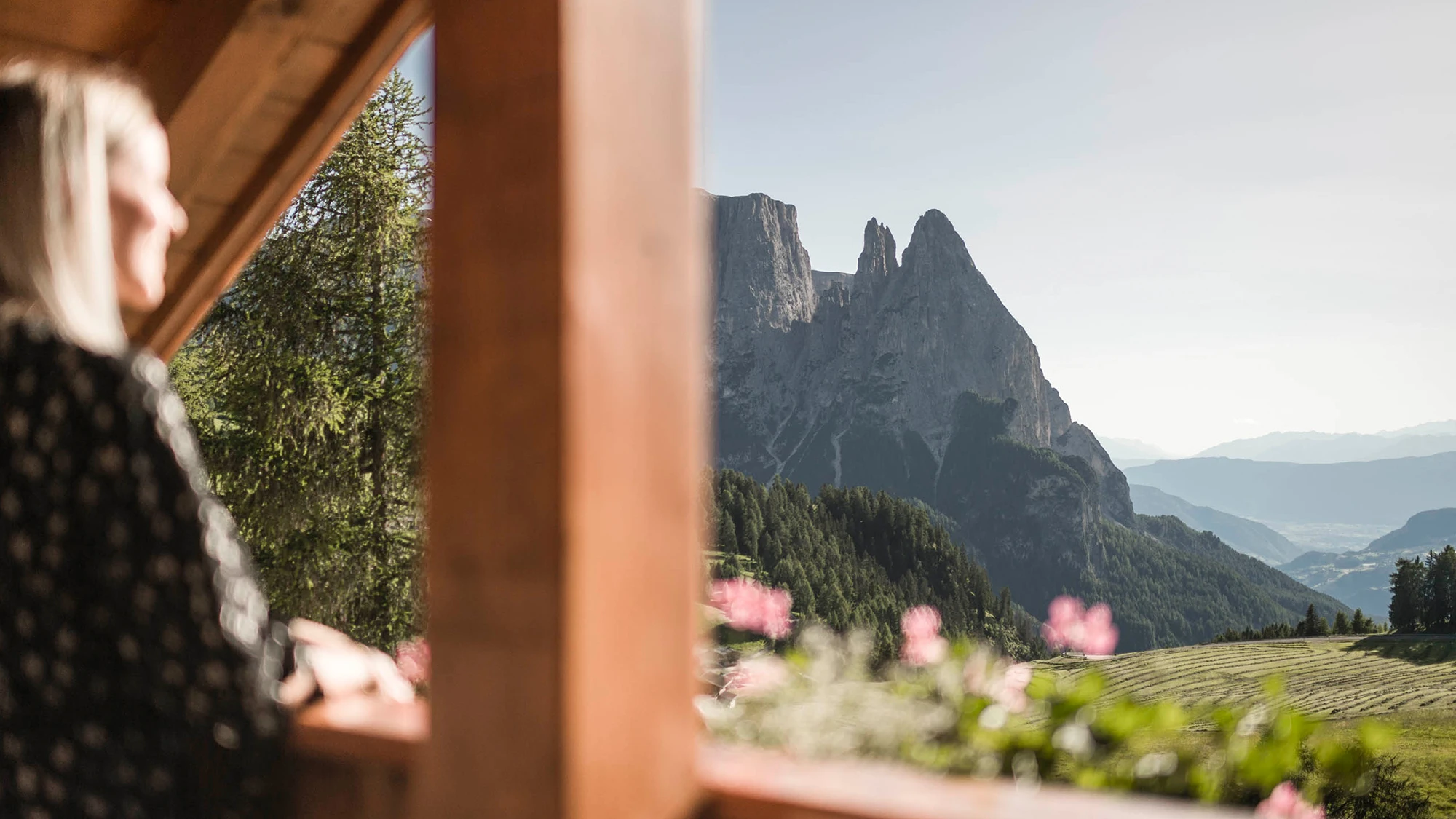 A woman gazes at the majestic Dolomites from a wooden balcony in bright sunshine.