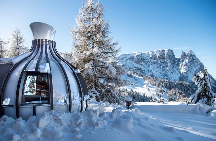 Una moderna cupola di vetro nella neve davanti all'imponente paesaggio montano delle Dolomiti sull'Alpe di Siusi, vicino all'Hotel Rosa durante le vostre vacanze invernali. - Bild größer darstellen