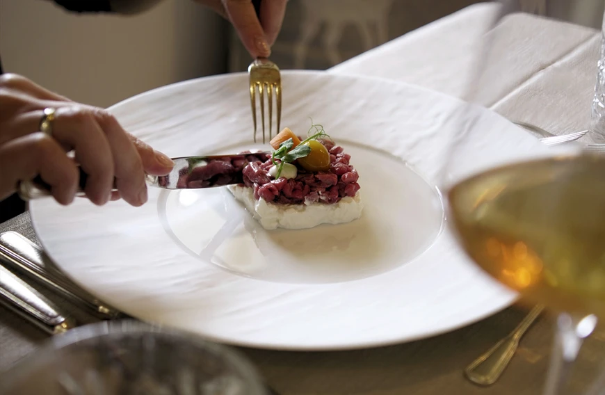 Una persona usa coltello e forchetta per tagliare una tartare di manzo preparata delicatamente su un piatto bianco in un elegante ristorante con cuscini di cervo. - Bild größer darstellen