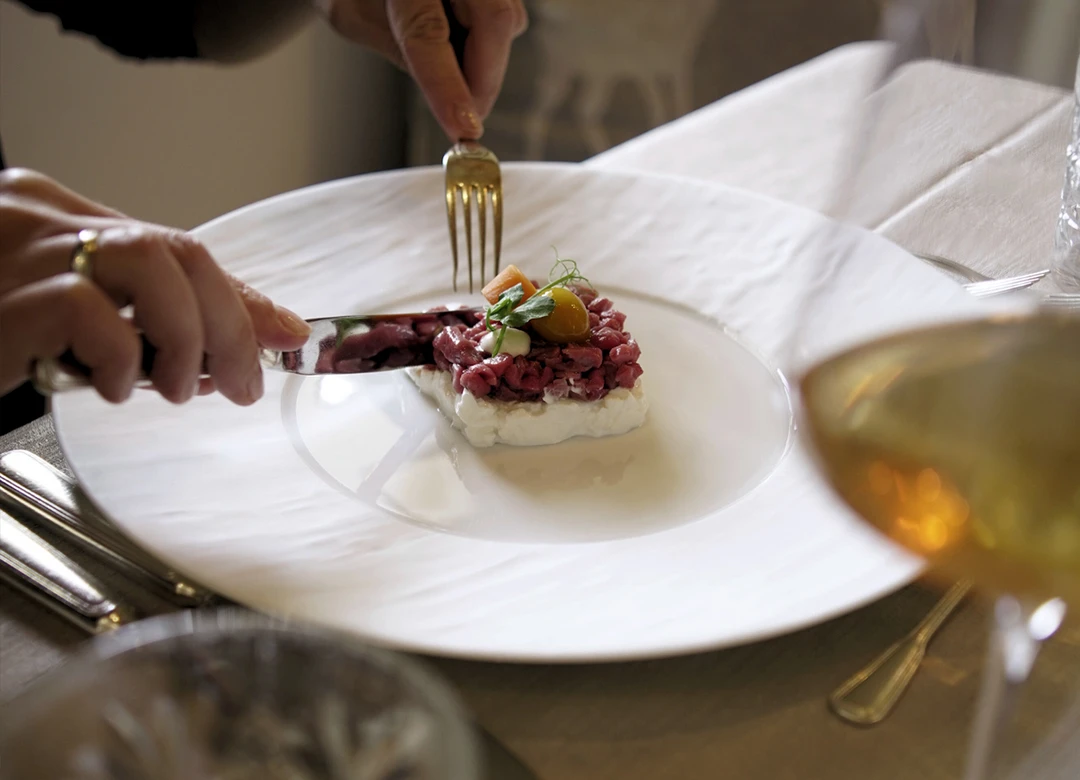 Una persona usa coltello e forchetta per tagliare una tartare di manzo preparata delicatamente su un piatto bianco in un elegante ristorante con cuscini di cervo. - Bild größer darstellen