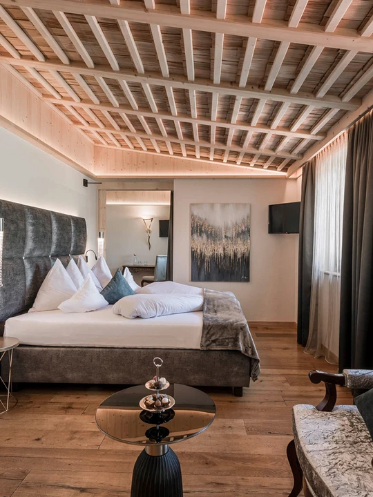 Una lussuosa camera d'albergo con un comodo letto, soffitto con travi in ​​legno, un'elegante poltrona e un bagno annesso con doppio lavabo. - Bild größer darstellen