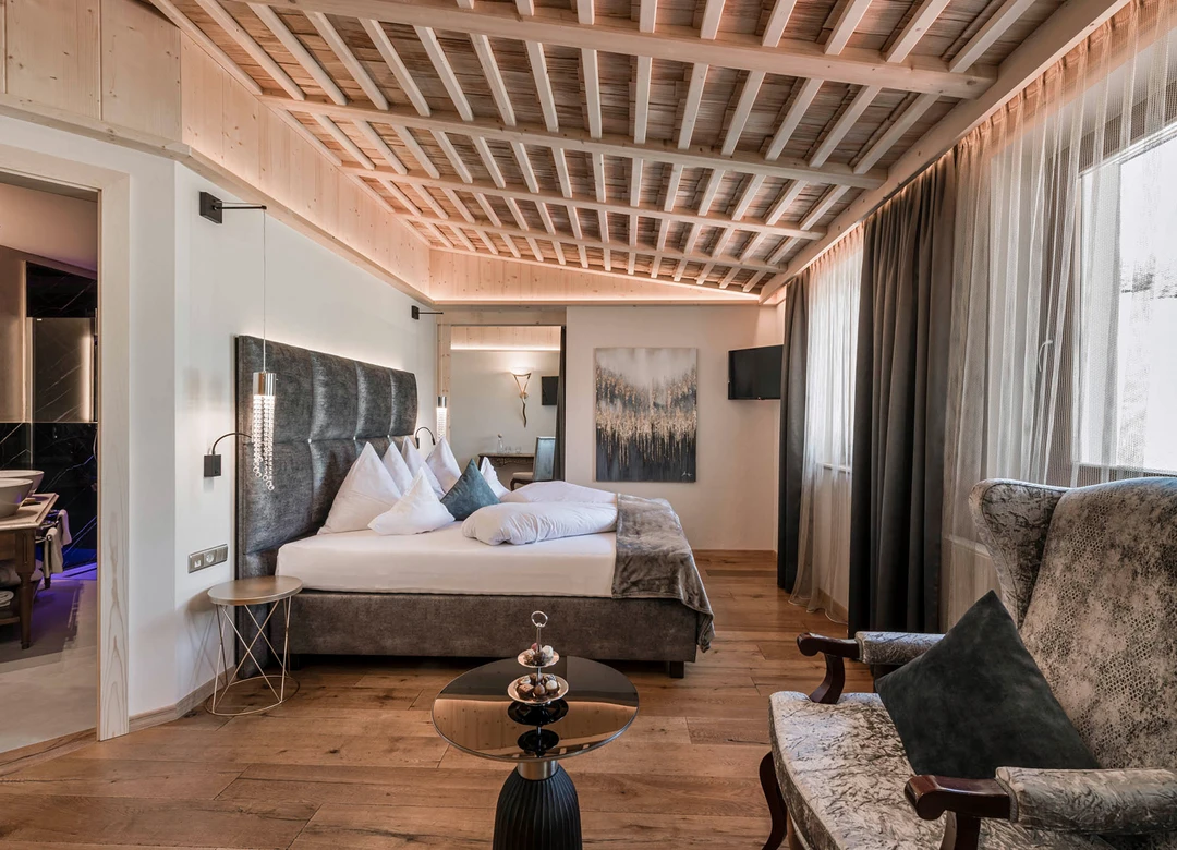 Una lussuosa camera d'albergo con un comodo letto, soffitto con travi in ​​legno, un'elegante poltrona e un bagno annesso con doppio lavabo. - Bild größer darstellen