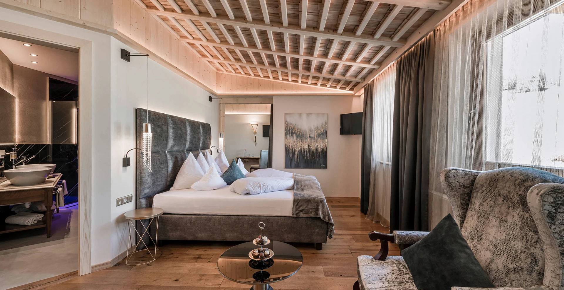 Una lussuosa camera d'albergo con un comodo letto, soffitto con travi in ​​legno, un'elegante poltrona e un bagno annesso con doppio lavabo.