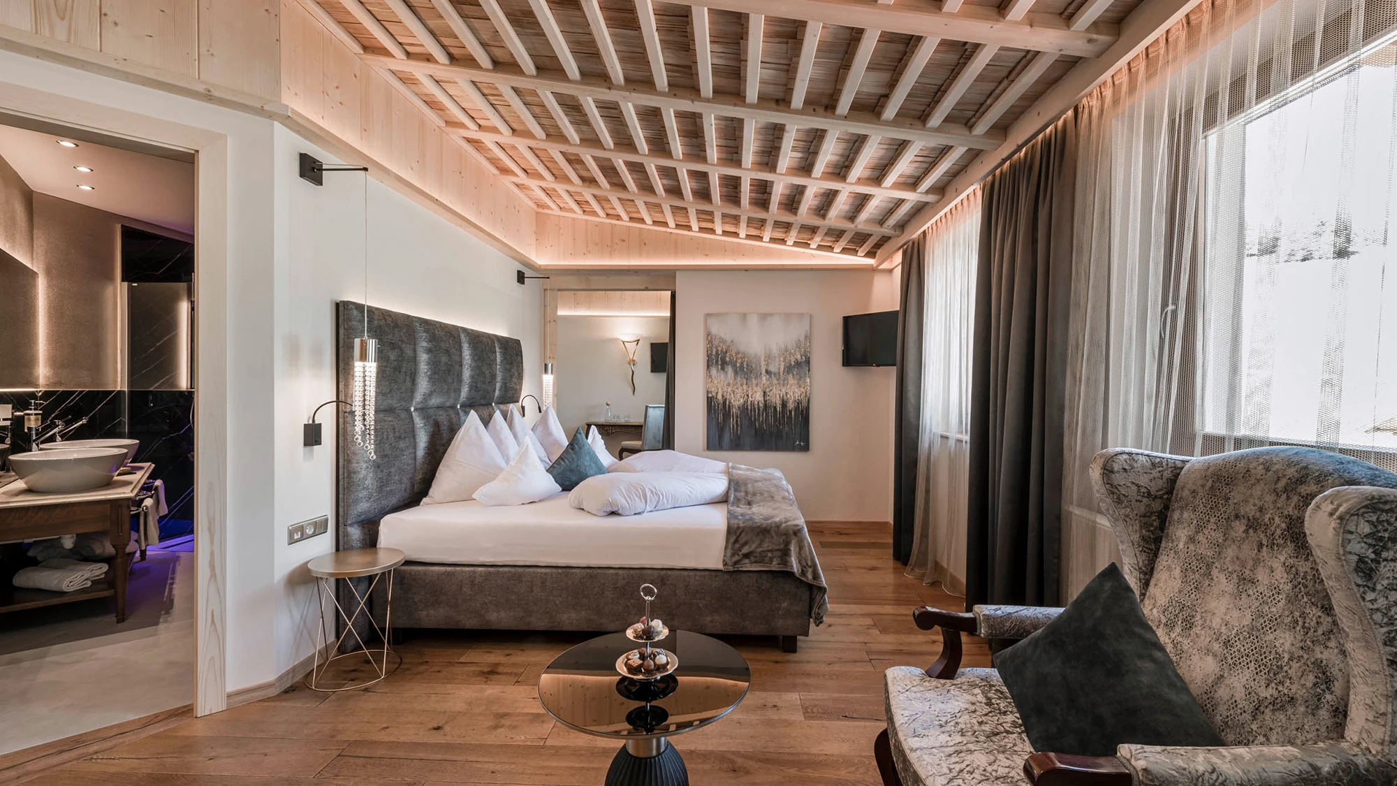 Una lussuosa camera d'albergo con un comodo letto, soffitto con travi in ​​legno, un'elegante poltrona e un bagno annesso con doppio lavabo.