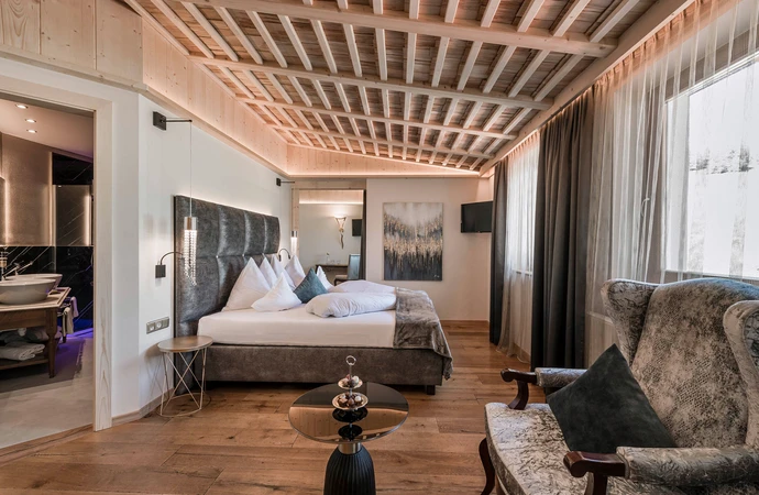 Una lussuosa camera d'albergo con un comodo letto, soffitto con travi in ​​legno, un'elegante poltrona e un bagno annesso con doppio lavabo. - Bild größer darstellen