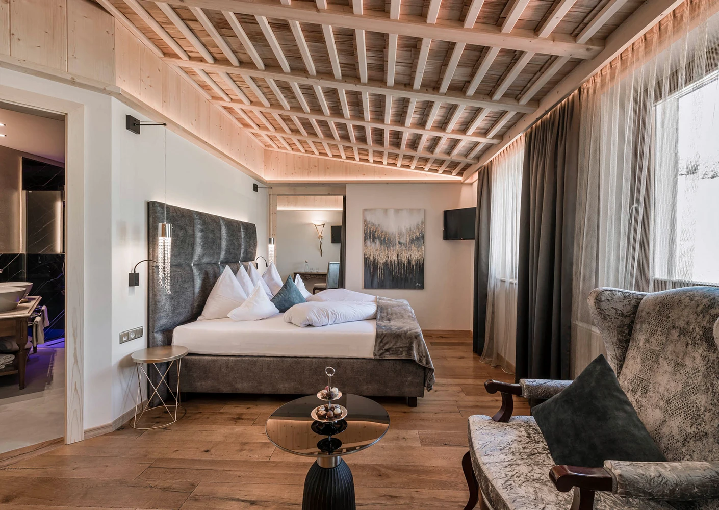 Una lussuosa camera d'albergo con un comodo letto, soffitto con travi in ​​legno, un'elegante poltrona e un bagno annesso con doppio lavabo.