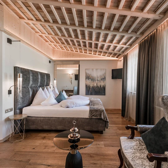 Una lussuosa camera d'albergo con un comodo letto, soffitto con travi in ​​legno, un'elegante poltrona e un bagno annesso con doppio lavabo.