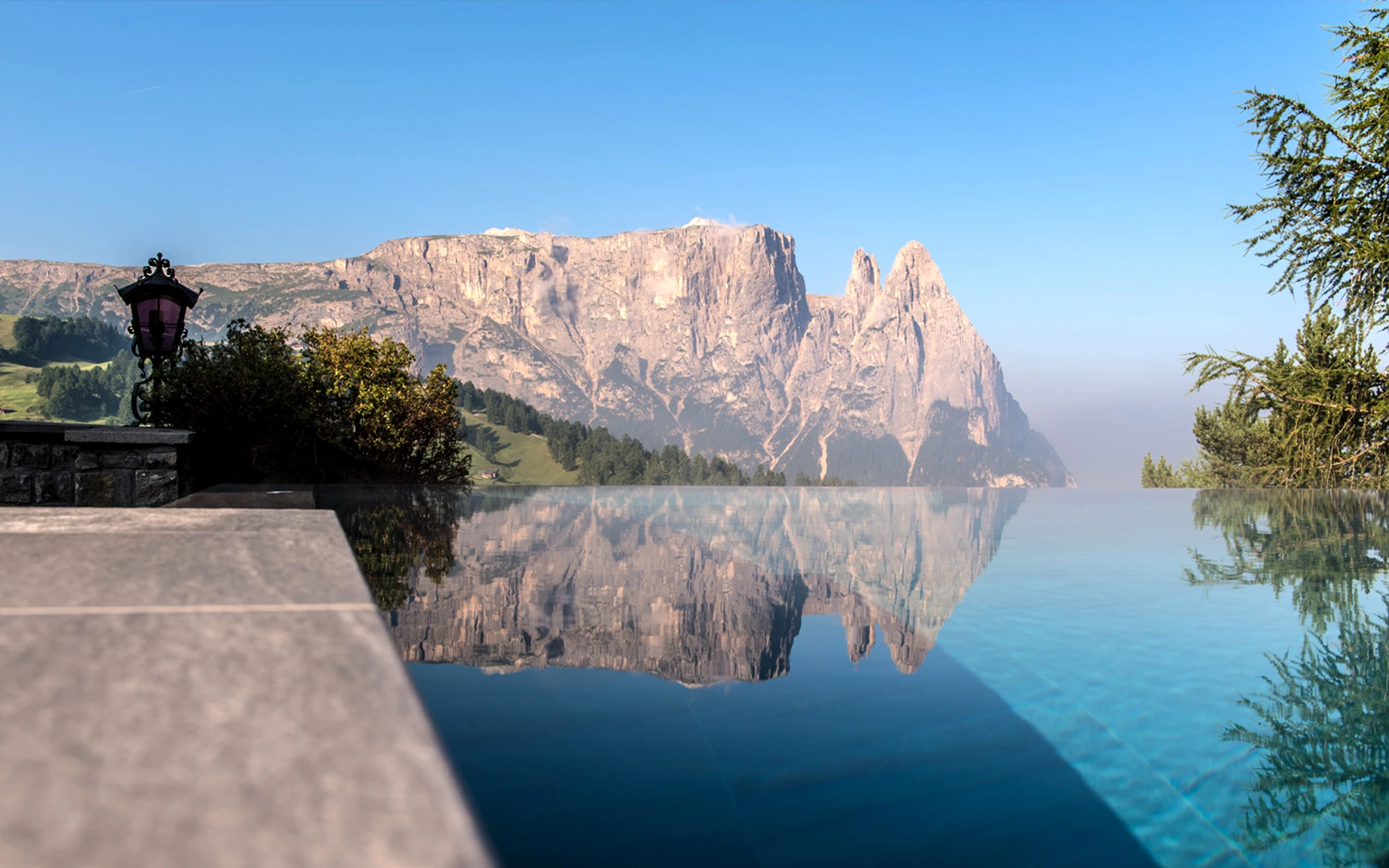 Una piscina infinity riflette l’imponente massiccio montuoso delle Dolomiti, sotto un cielo di un blu intenso e in una splendida giornata di sole.