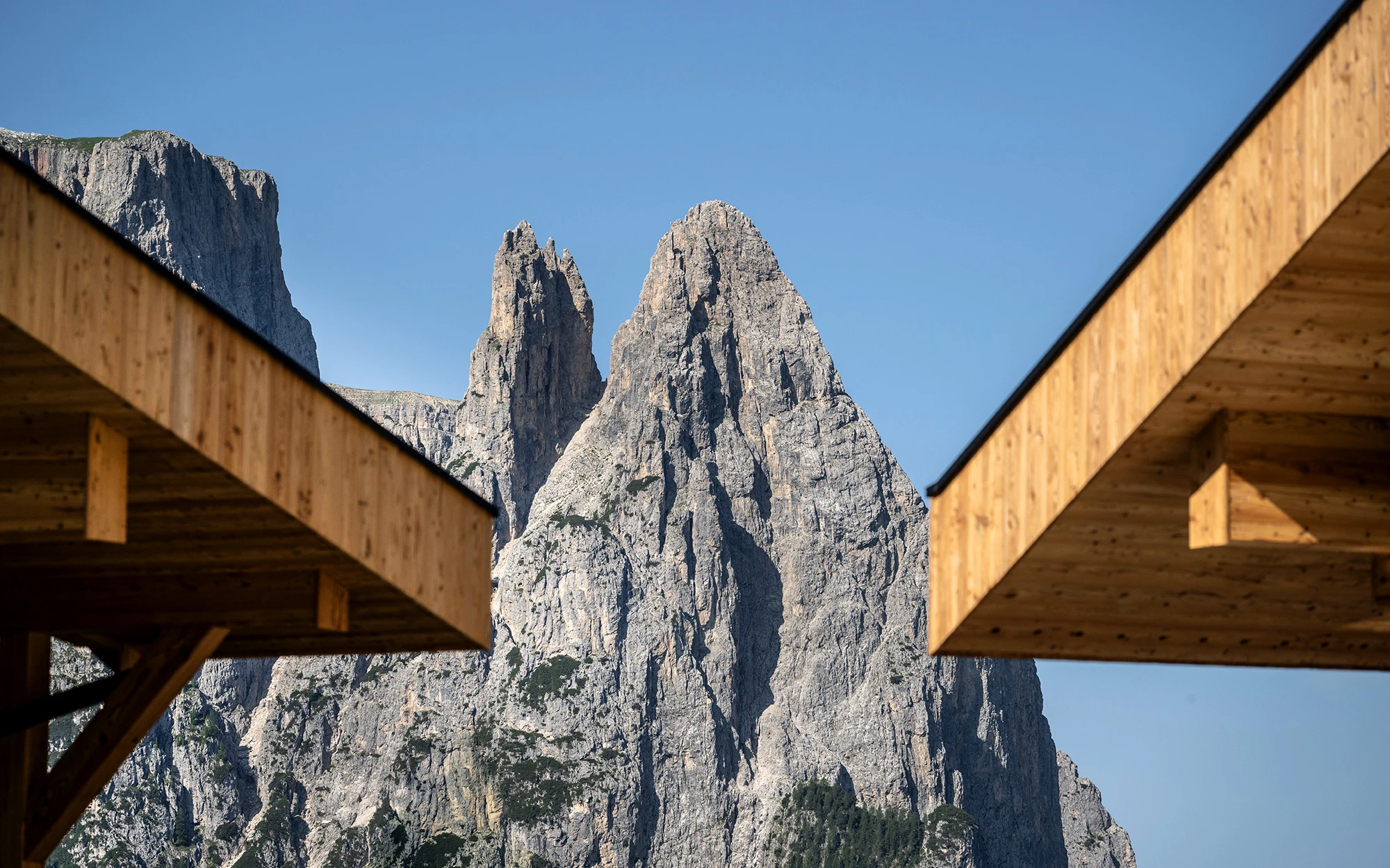 Vista attraverso due grondaie del tetto in legno sulle imponenti e aspre formazioni rocciose delle Dolomiti sotto un cielo azzurro.
