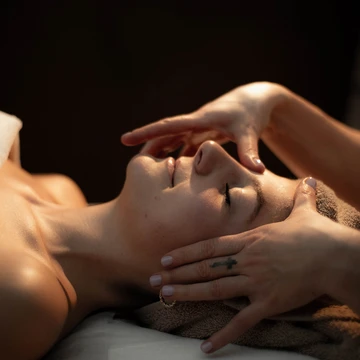 Una giovane donna a occhi chiusi si gode un benefico massaggio al viso in un’atmosfera rilassante e dalle luci soffuse. - Bild größer darstellen